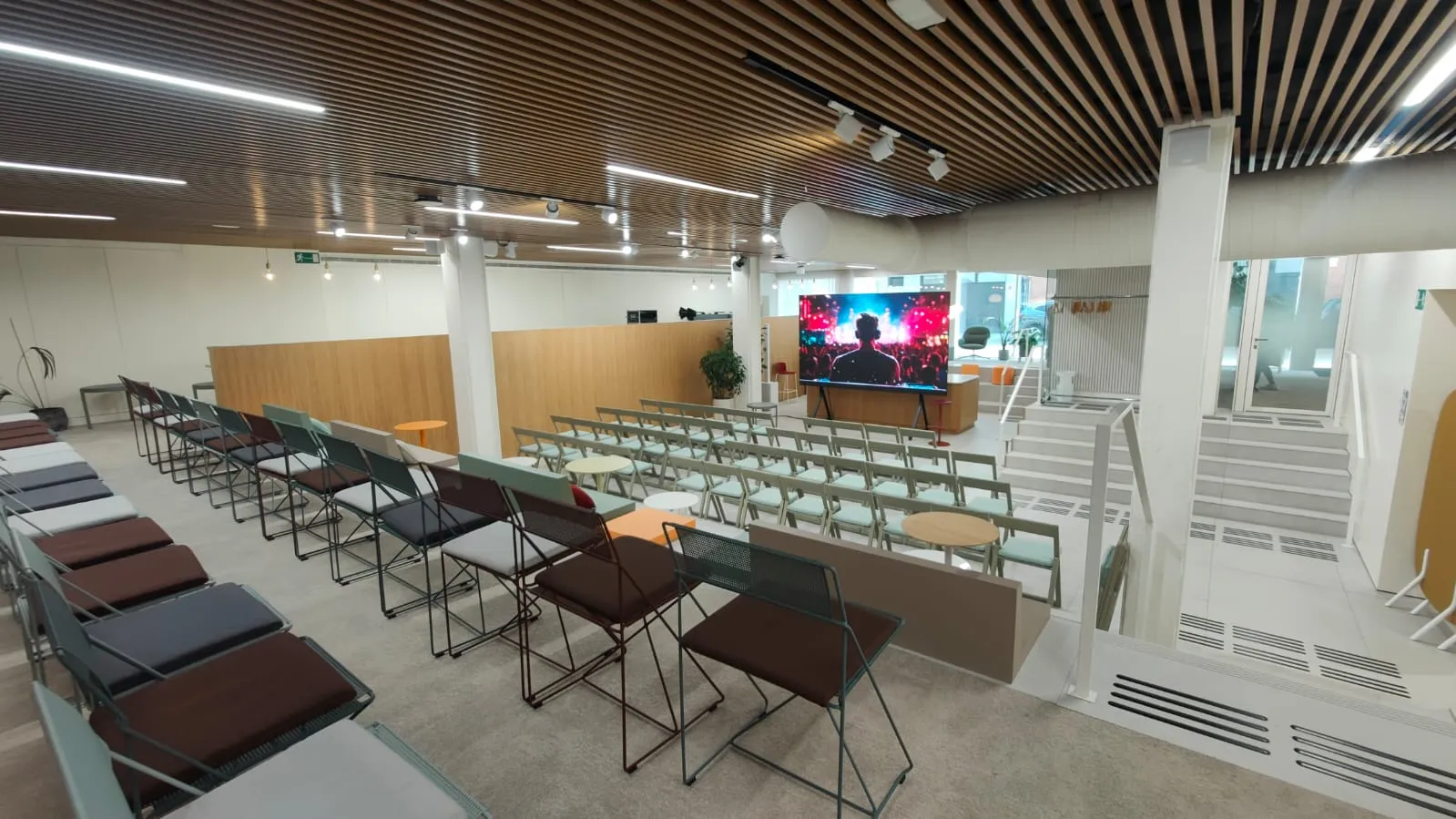 Sala Ágora con filas de sillas frente a una gran pantalla LED, techo de lamas de madera y escalera lateral